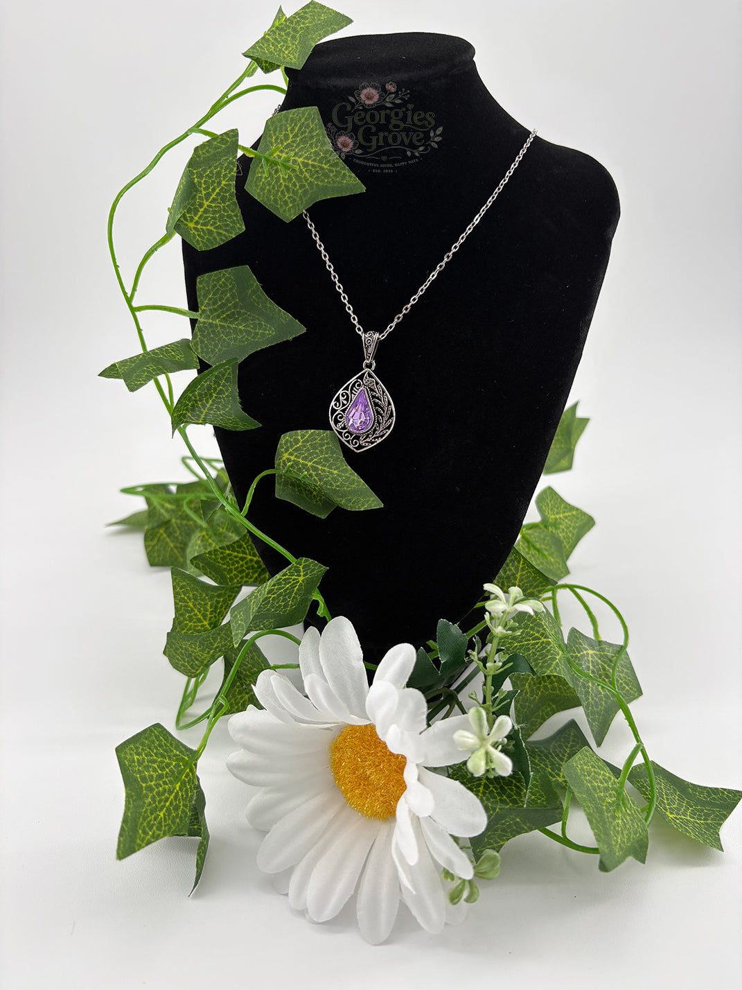 Necklace with a purple pendant on a black mannequin surrounded by green leaves and a white flower.