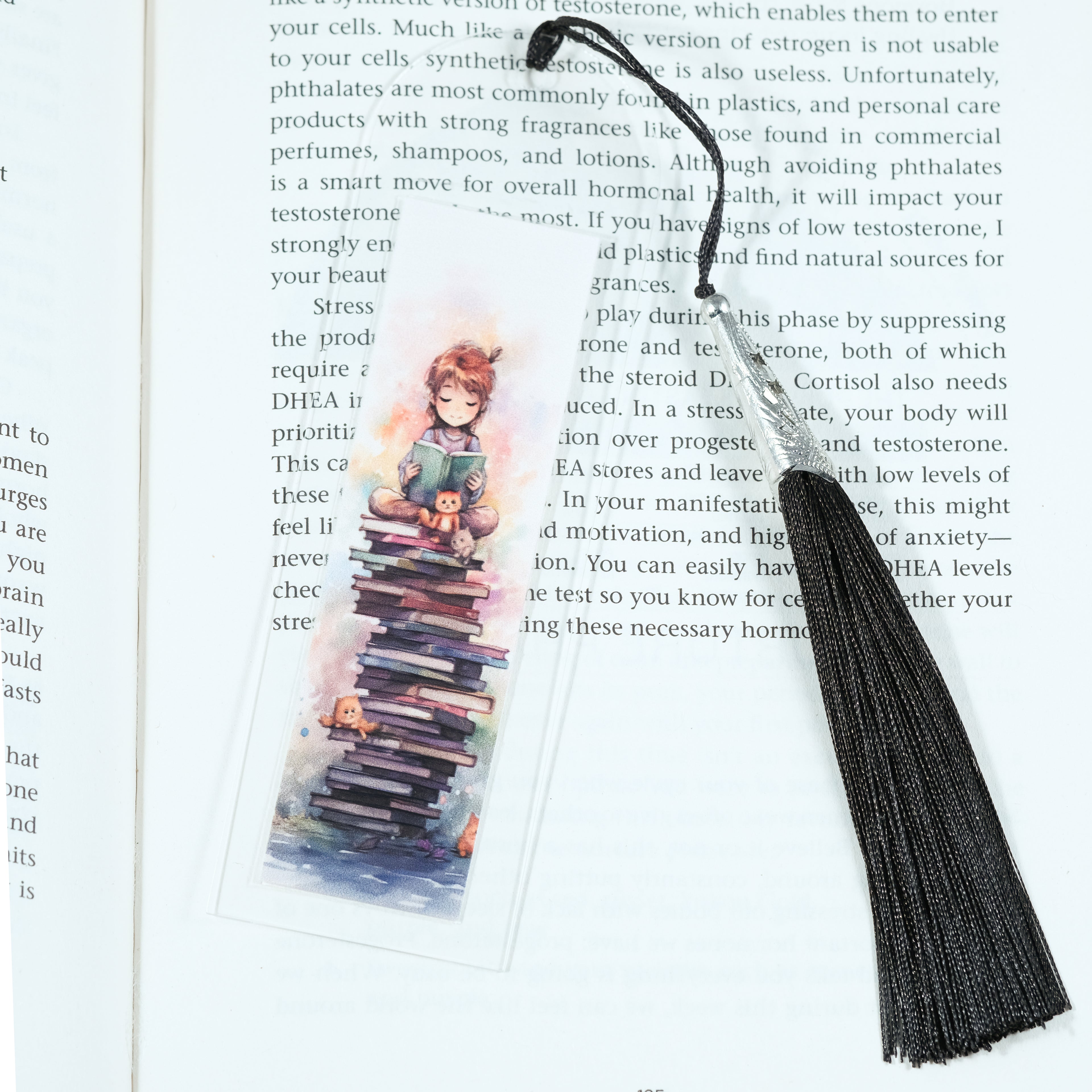 Mark your place in style! This cheerful acrylic bookmark features a delightful design of a young girl sitting on a tower of books reading to her companions celebrating the joy of reading.