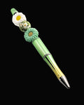 A graduated green to gold metallic pen with a green and a white daisy shaped beads and a round bead with a daisy pattern.