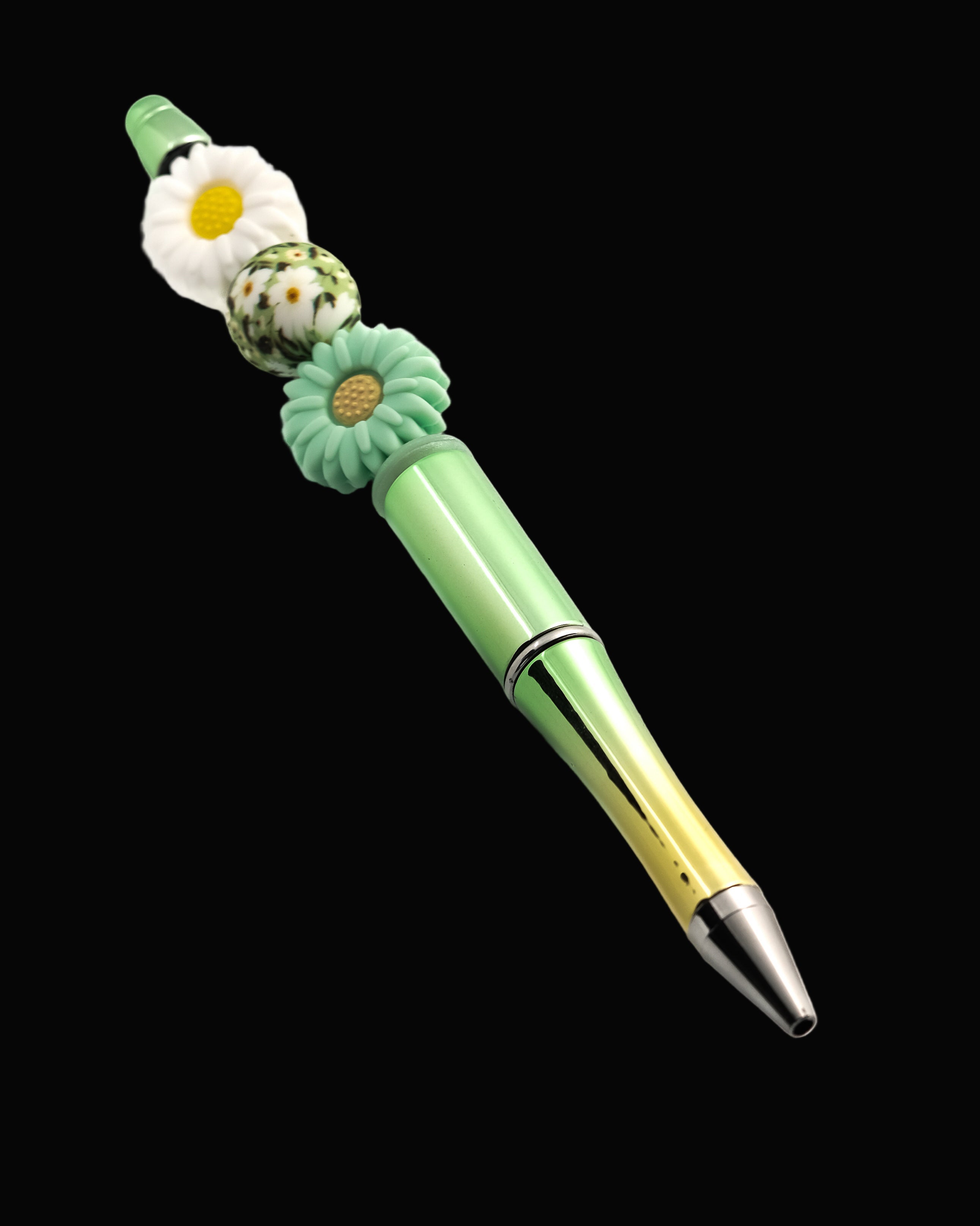 A graduated green to gold metallic pen with a green and a white daisy shaped beads and a round bead with a daisy pattern.