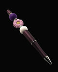 A purple metallic pen with a purple daisy shaped bead, a purple round bead, a white bead and sparkly Rondelle.