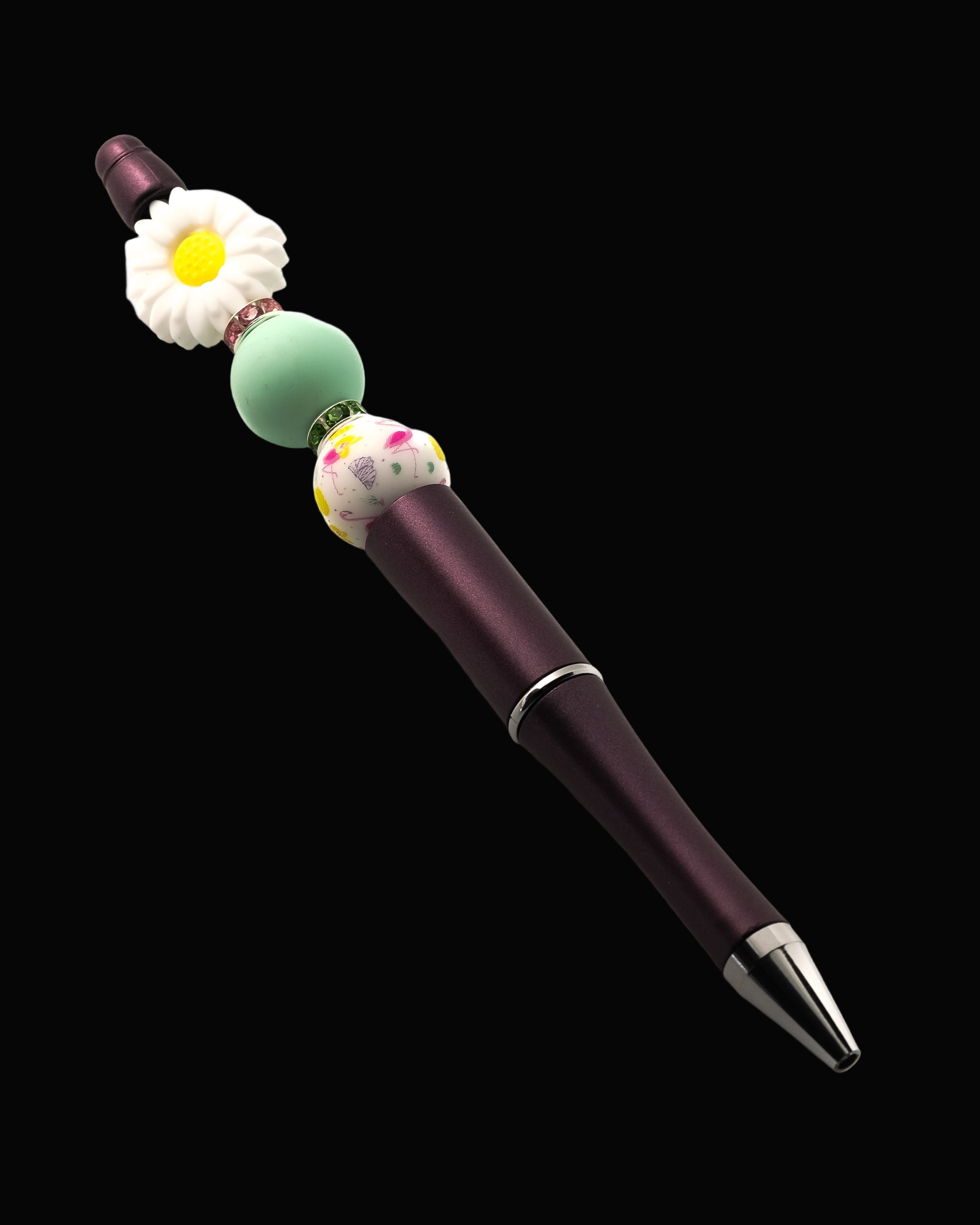 A purple metallic pen with a white daisy shaped bead, a green round bead, a white bead with a floral design and a pink and a green Rondelle.