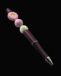 A purple metallic pen with a purple daisy shaped bead, a purple round bead, a white bead with a floral design and a pink and a green Rondelle.